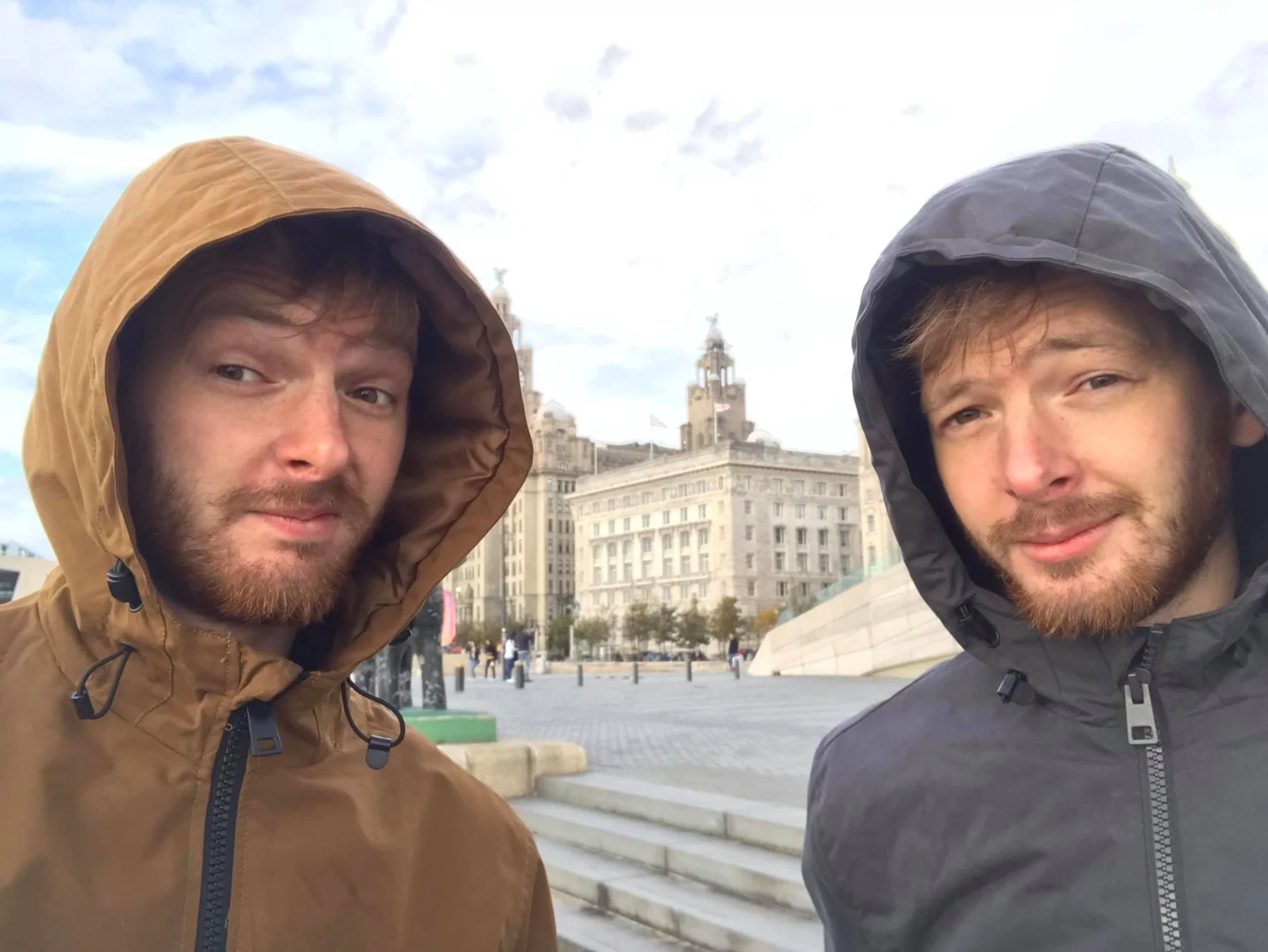 Two young men wearing hooded jackets taking a selfie outdoors in front of a historic building with clock towers and flagpoles, under a cloudy sky.