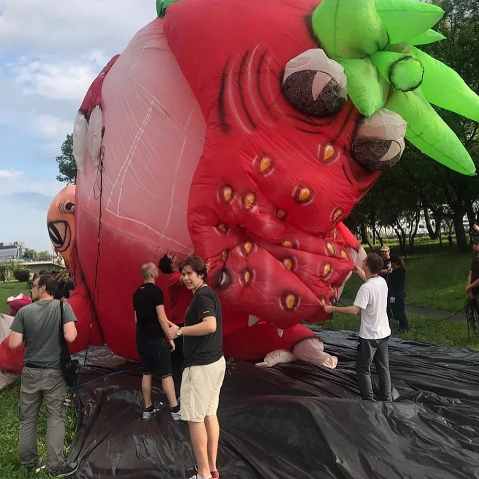 People setting up a large inflatable strawberry with green leaves and big eyes outdoors on a grassy area.