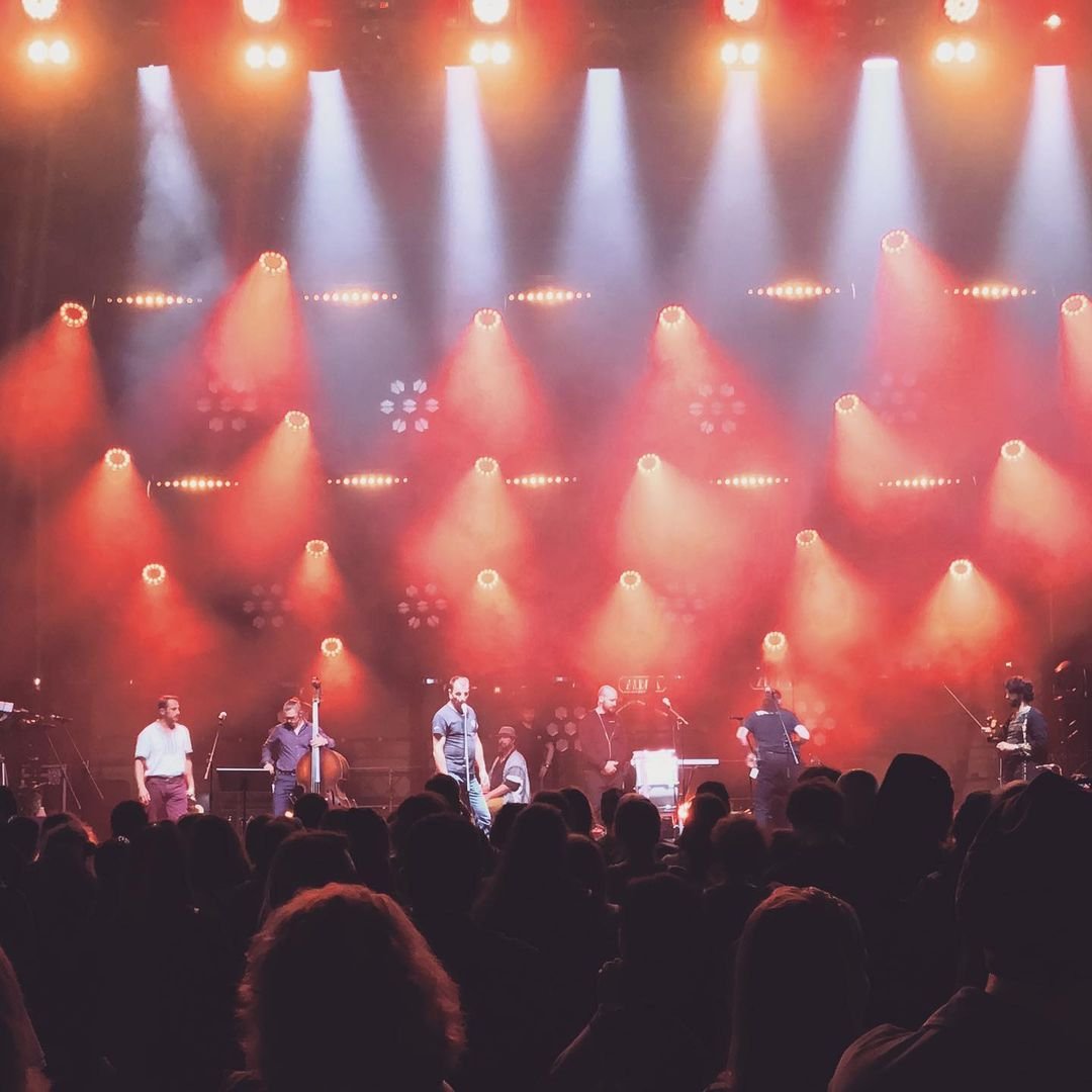 Musicians performing on stage under red and orange stage lights with an audience watching.