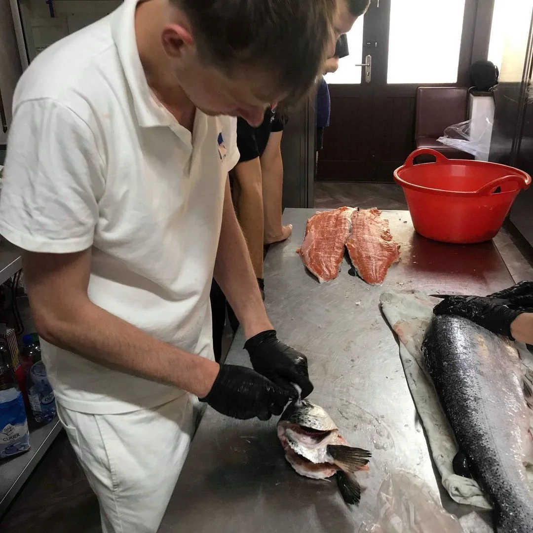 A person wearing white attire and black gloves preparing fish on a stainless steel counter. Fish fillets and a whole fish are visible on the counter, with a red bucket nearby.