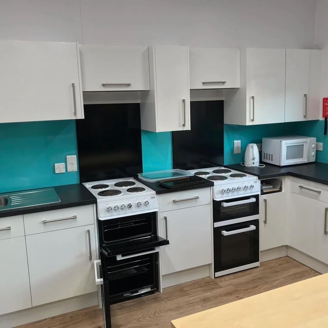 Kitchen with white cabinets, teal backsplash, black countertops, electric stove, oven, microwave, kettle, and wooden floor.