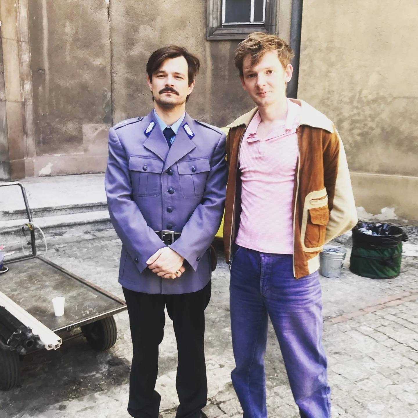 Two men standing outdoors against a concrete building wall, one in a police uniform and the other in casual clothing.