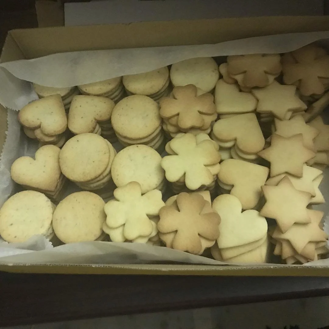 A box of assorted cookies shaped like hearts, stars, clouds, and flowers, with some stacked inside a cardboard box lined with paper.