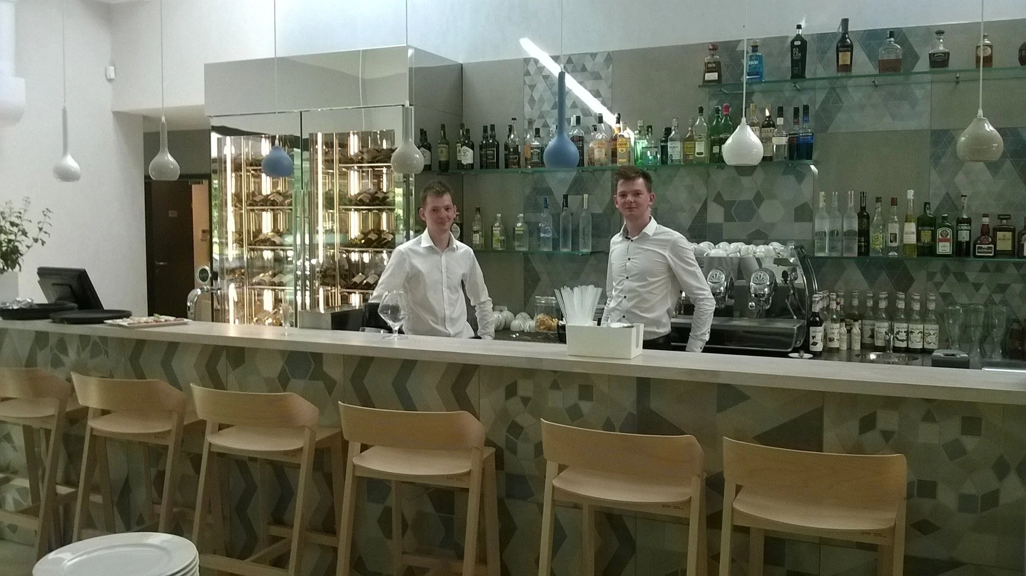 Two young men in white shirts standing behind a bar counter in a modern, stylish bar or restaurant. The bar features a geometric-patterned front, with shelves of liquor bottles and a glass display of pastries or bread. There are colorful hanging pend