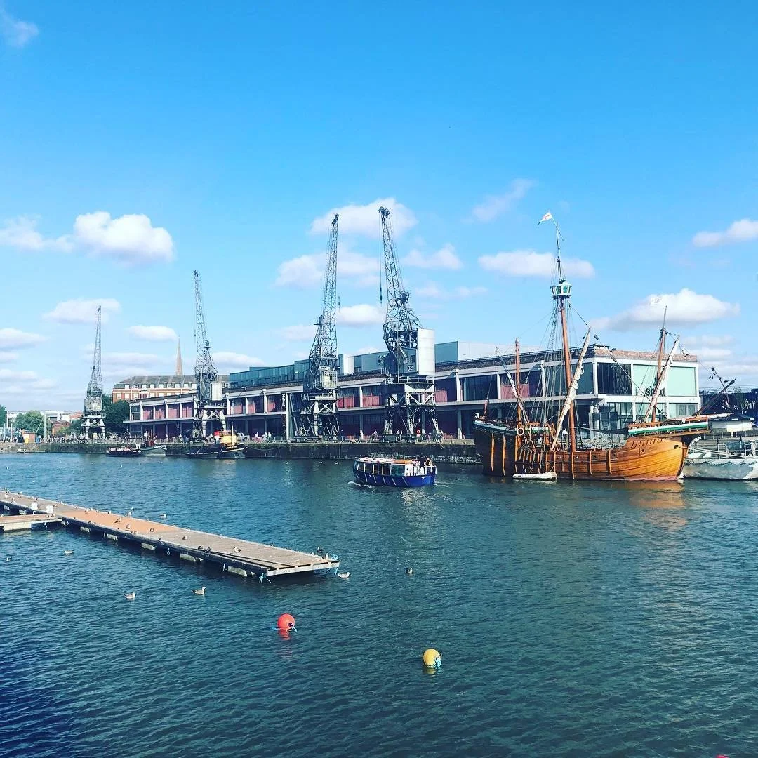 A harbor with modern building along the waterfront, two large cranes, a historical wooden ship, and several smaller boats on calm water under a blue sky with scattered clouds.