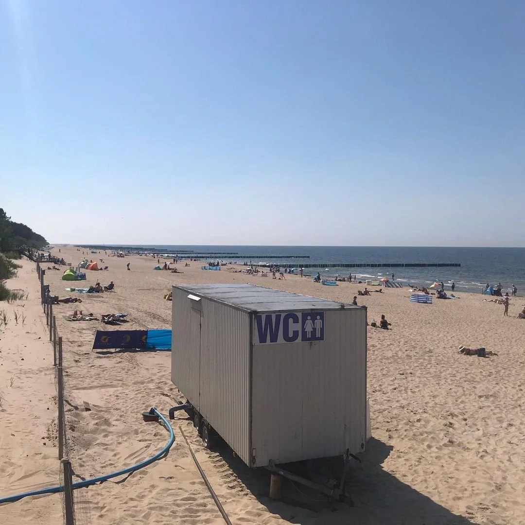Beach scene with people relaxing and umbrellas, a portable restroom trailer marked "WC," and ocean with a dock or pier, under clear blue sky.