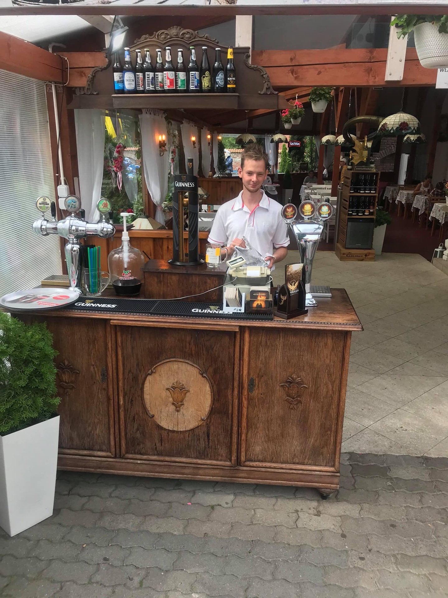 A young man stands behind a wooden bar counter inside a restaurant, with beer taps and bottles on display above him. The restaurant's interior features tables with white tablecloths and hanging plants.