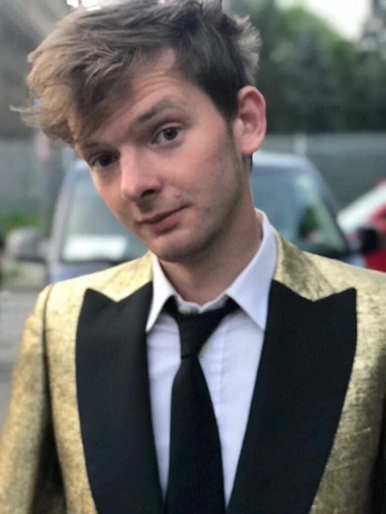 A young man with brown hair wearing a tuxedo with gold shoulders and a black tie, standing outdoors in front of parked cars.