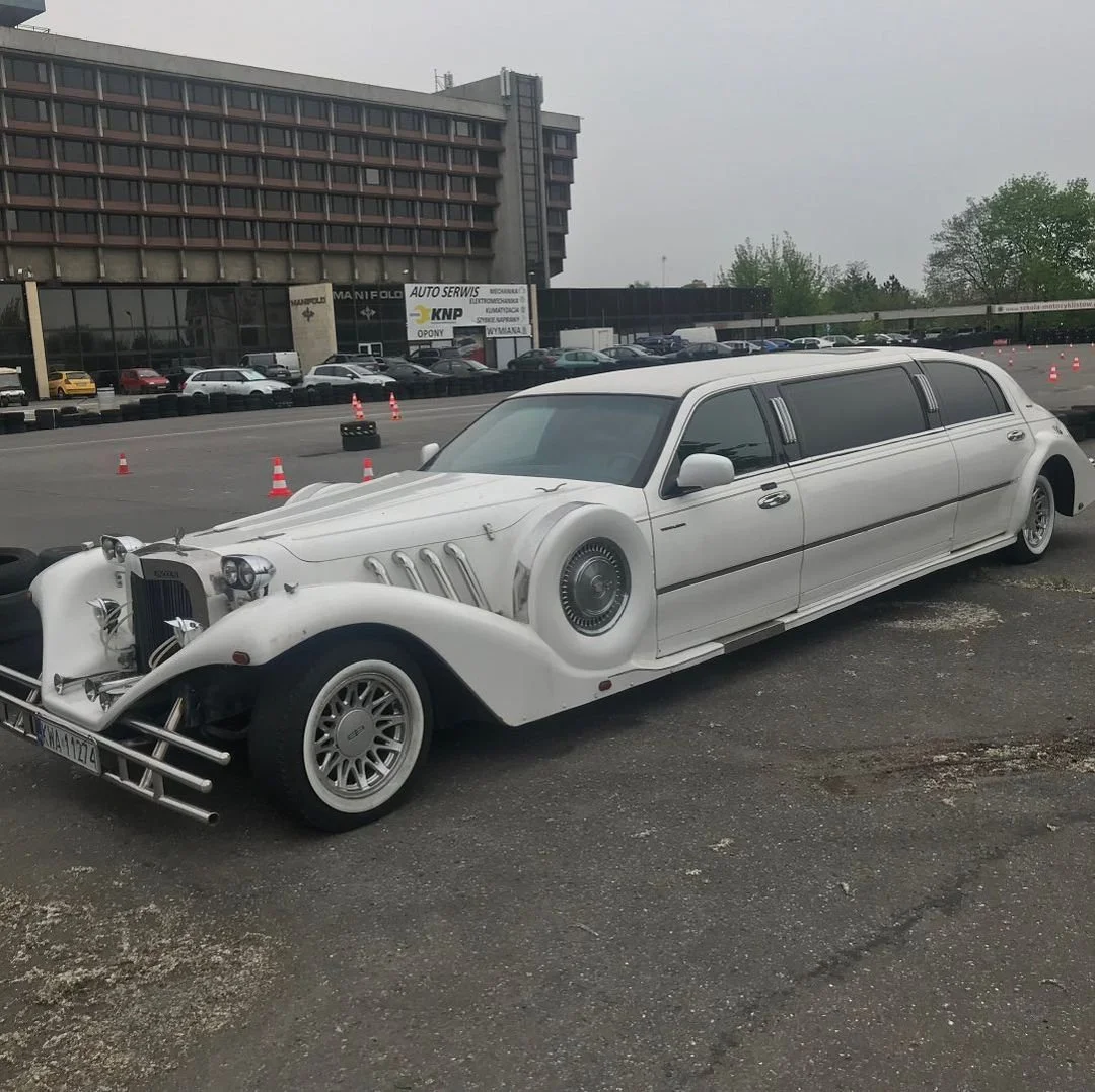 A white vintage limousine with customization resembling a classic car, parked on a lot near a building with an auto service shop, with other cars and parking lot cones in the background.