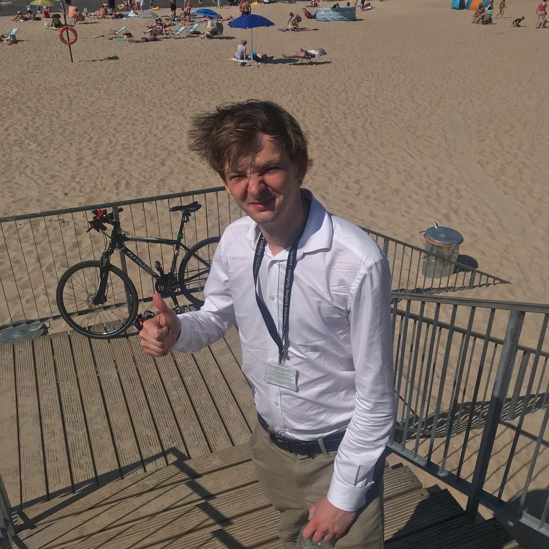 A young man with tousled hair waving and giving a thumbs-up at the beach, with a bicycle behind him on a railing and people sunbathing under umbrellas in the background.