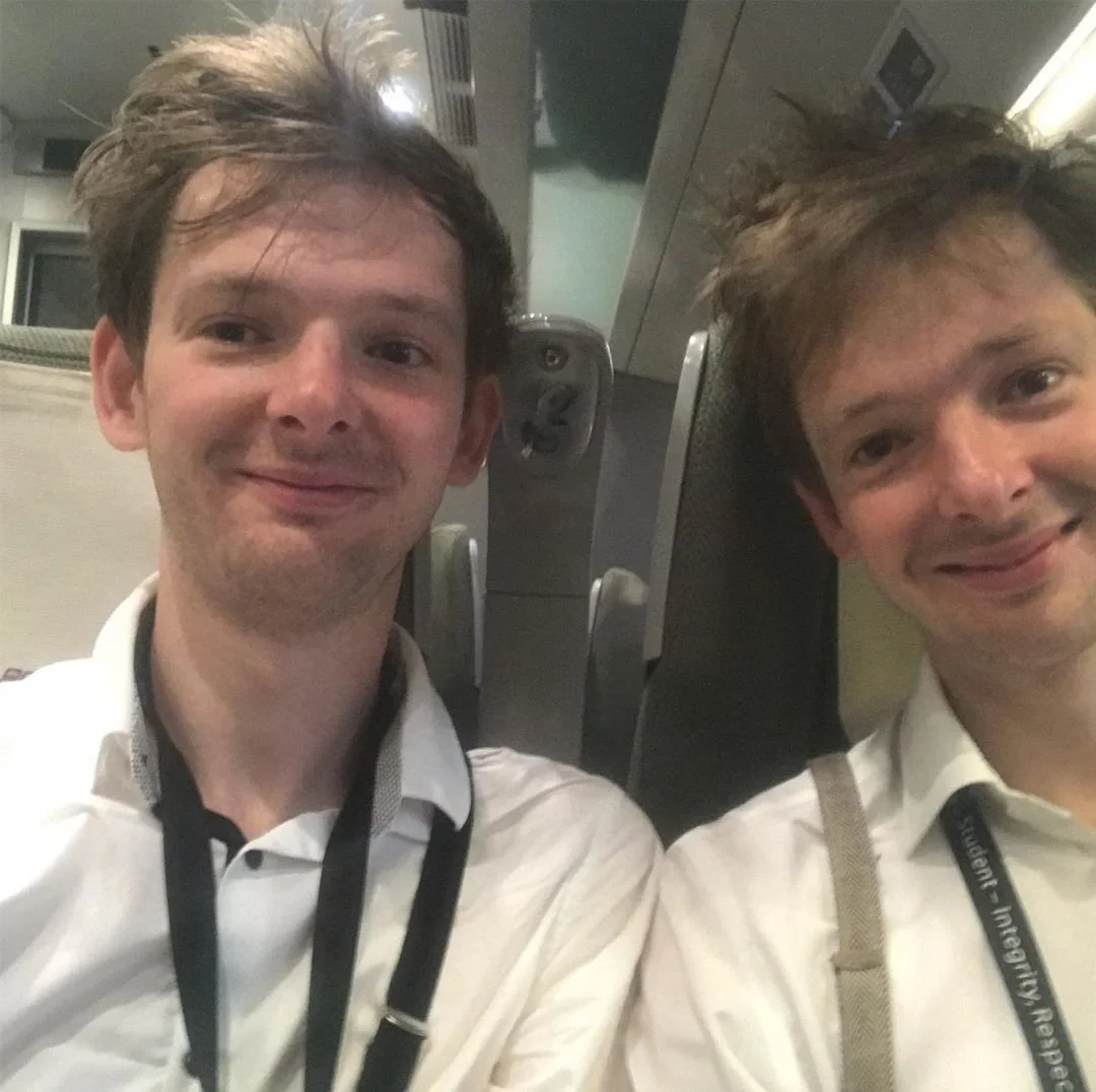 Two young men with tousled brown hair and white collared shirts taking a selfie, with seats and technology in the background.