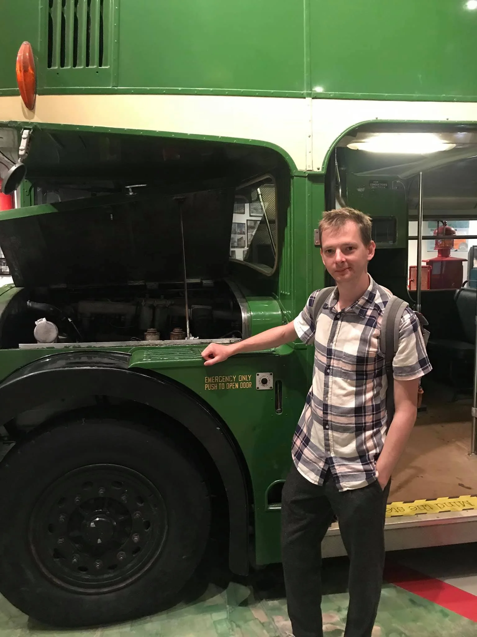 A young man in a plaid shirt and backpack standing next to a green double-decker bus with the hood open, showing the engine.