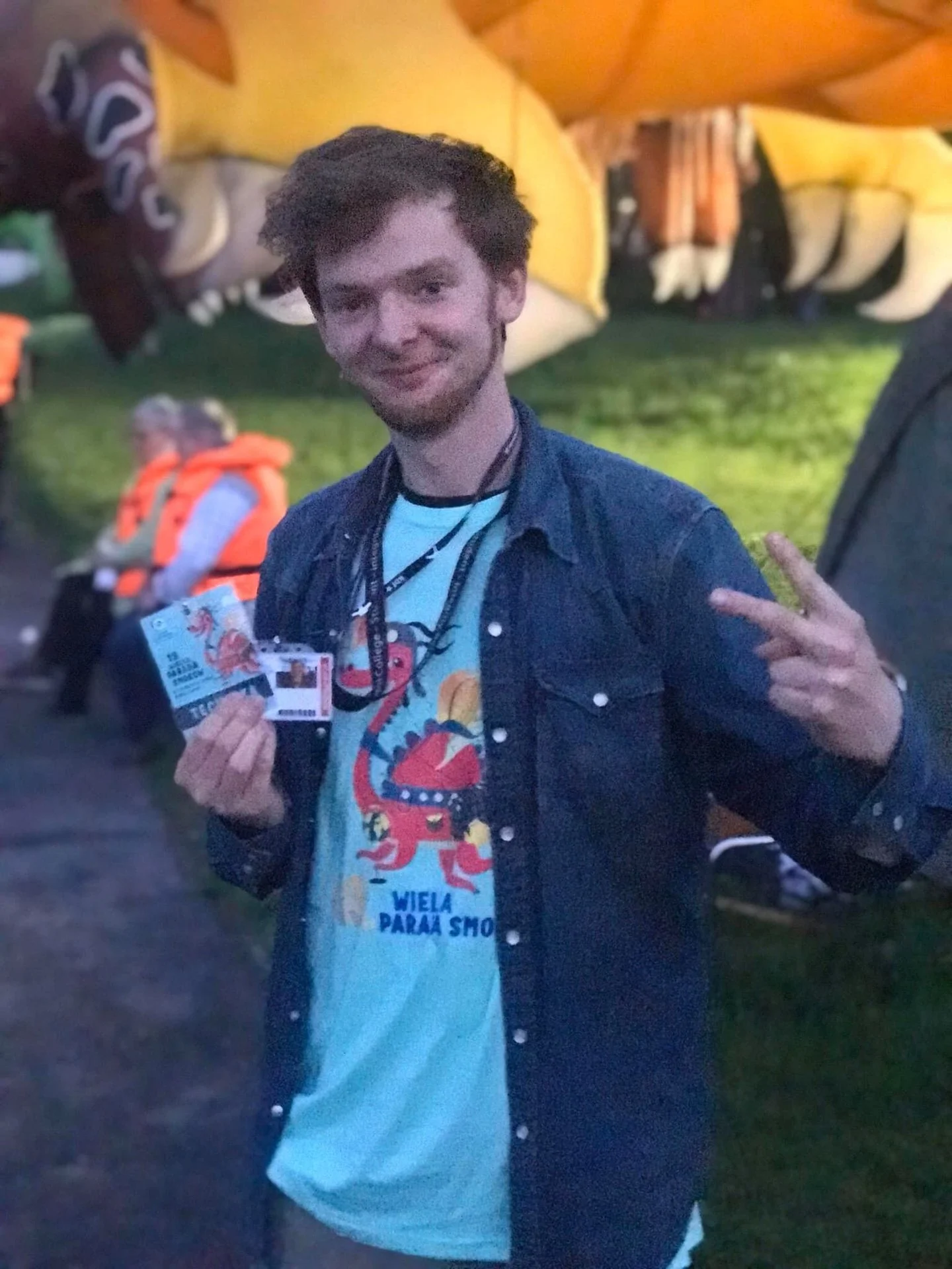 A young man with dark, curly hair and a beard is smiling and making a peace sign with his right hand. He is holding travel documents in his left hand and is wearing a light blue T-shirt with a red dragon graphic and the words "WIELA PARAŠ SHO" undern