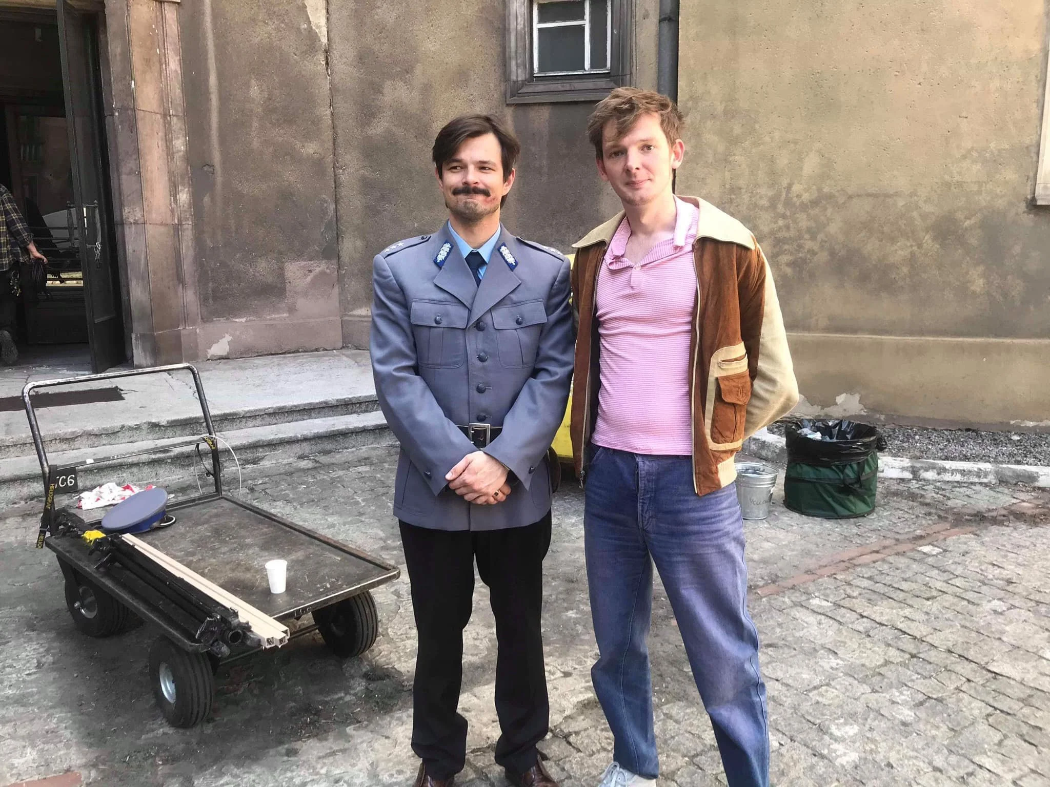 Two men standing on a cobblestone street in front of a plain wall, one dressed in a police uniform and the other in casual clothes, with a wheeled cart and some bags nearby.