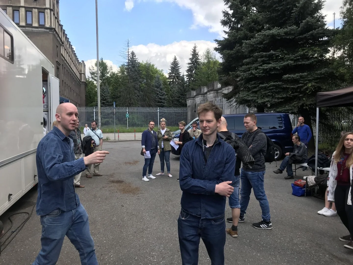 Group of people gathered outdoors near a mobile command unit with trees and a fence in the background. One man in the foreground is talking, and others are standing or sitting nearby.