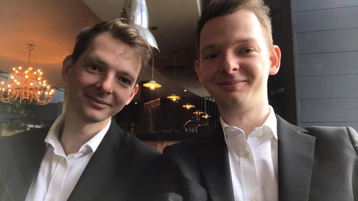 Two young men in formal attire taking a selfie inside a restaurant or cafe with warm lighting and chandelier decor.