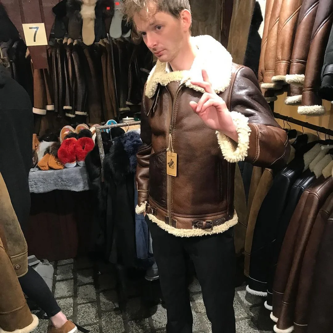 A young man with curly blond hair trying on a brown leather jacket with white shearling lining in a clothing store. He is making a peace sign with his right hand while looking to the side. The store has jackets hanging on walls and shelves.