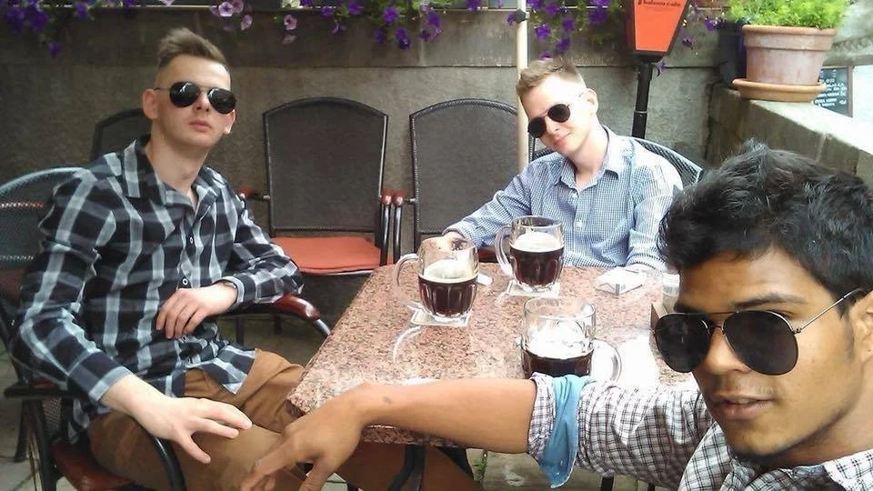 Three young men sitting at an outdoor table with four glasses of dark drinks, wearing sunglasses, on a patio with purple flowers and a concrete wall.