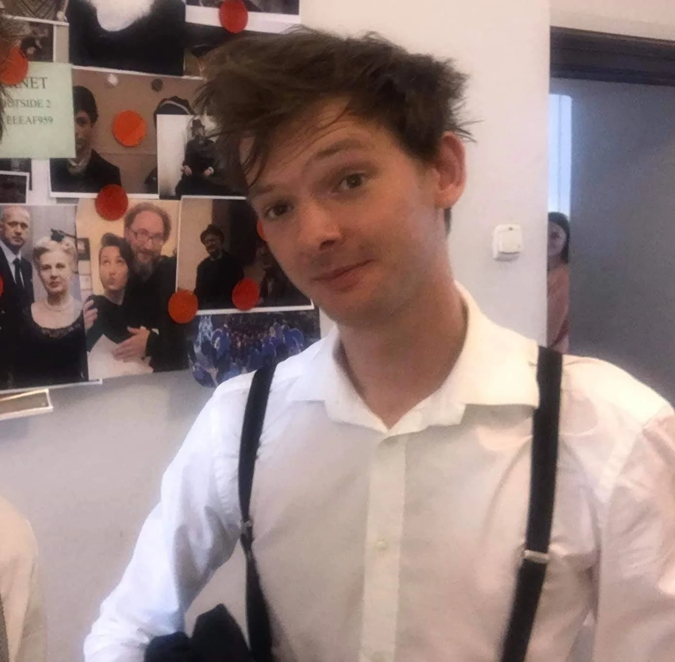 Young man with tousled brown hair wearing a white shirt and suspenders, standing in front of a photo collage on the wall.