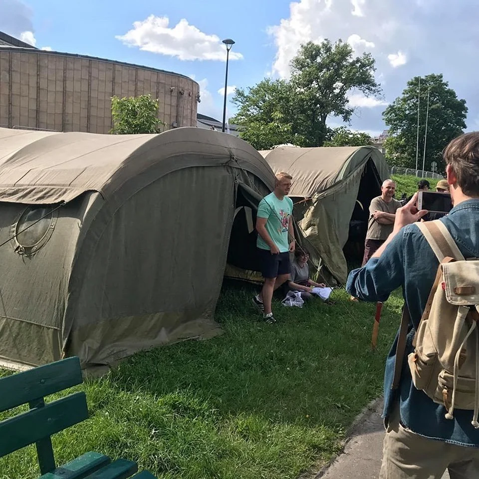 People taking photos of a group standing outside tents on a grassy area on a partly cloudy day.