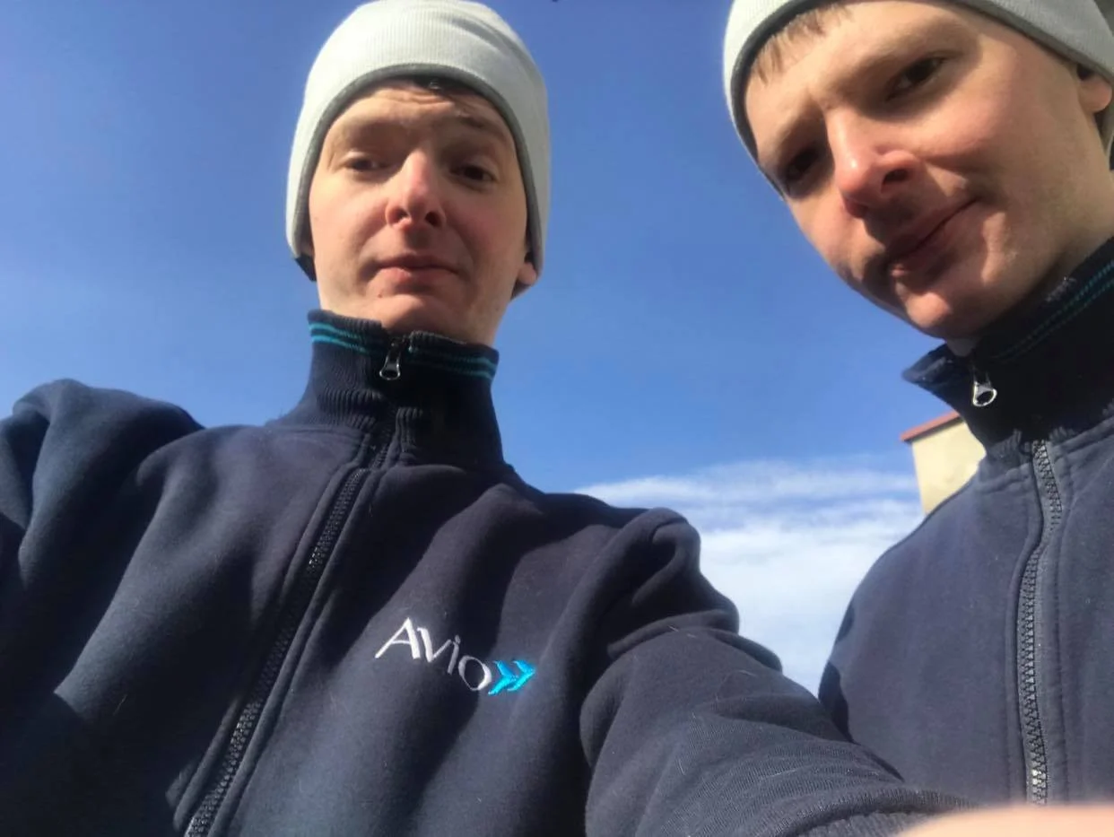 Two young men standing outdoors in jackets and beanies, taking a selfie against a clear blue sky.