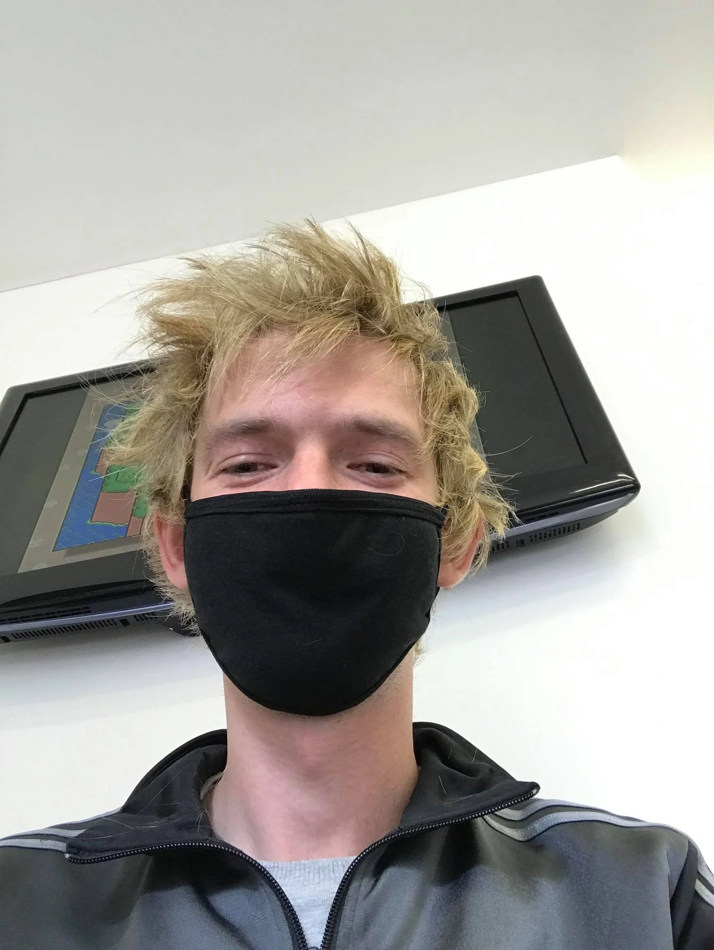 A young man with messy blonde hair wearing a black face mask and a black jacket, taking a selfie indoors with a laptop behind him.