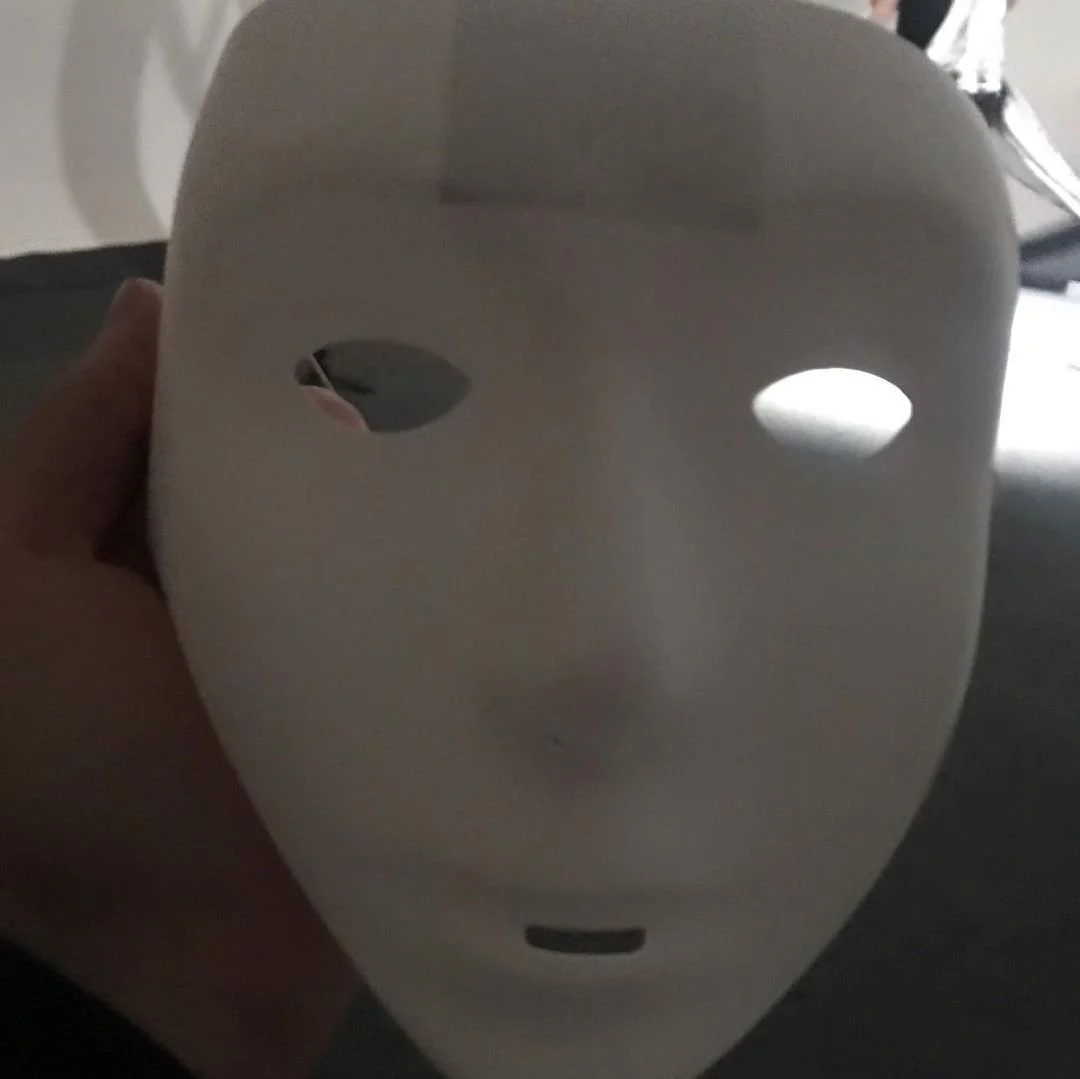 Person holding a white mask with holes for eyes and a small slit for a mouth, in a dimly lit room.