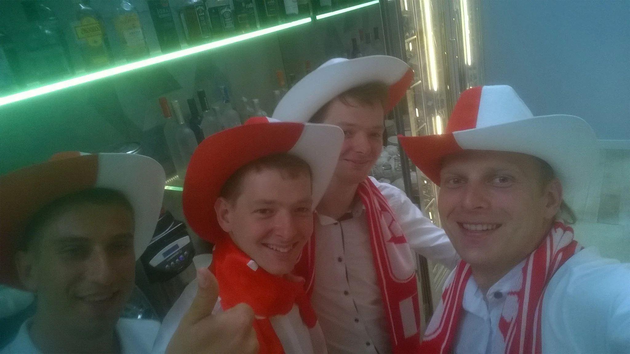 Four men wearing white hats with red accents, smiling, and dressed in white shirts with red scarves, posing for a selfie inside a store or bar with bottles on shelves behind them.