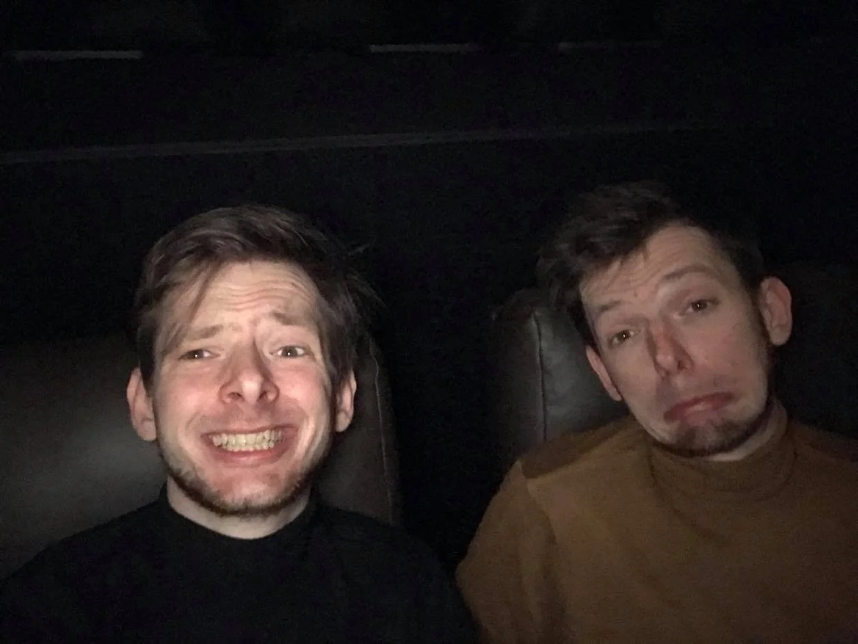 Two men sitting in a dark environment, one smiling with visible teeth and the other making a sad face, both facing the camera.