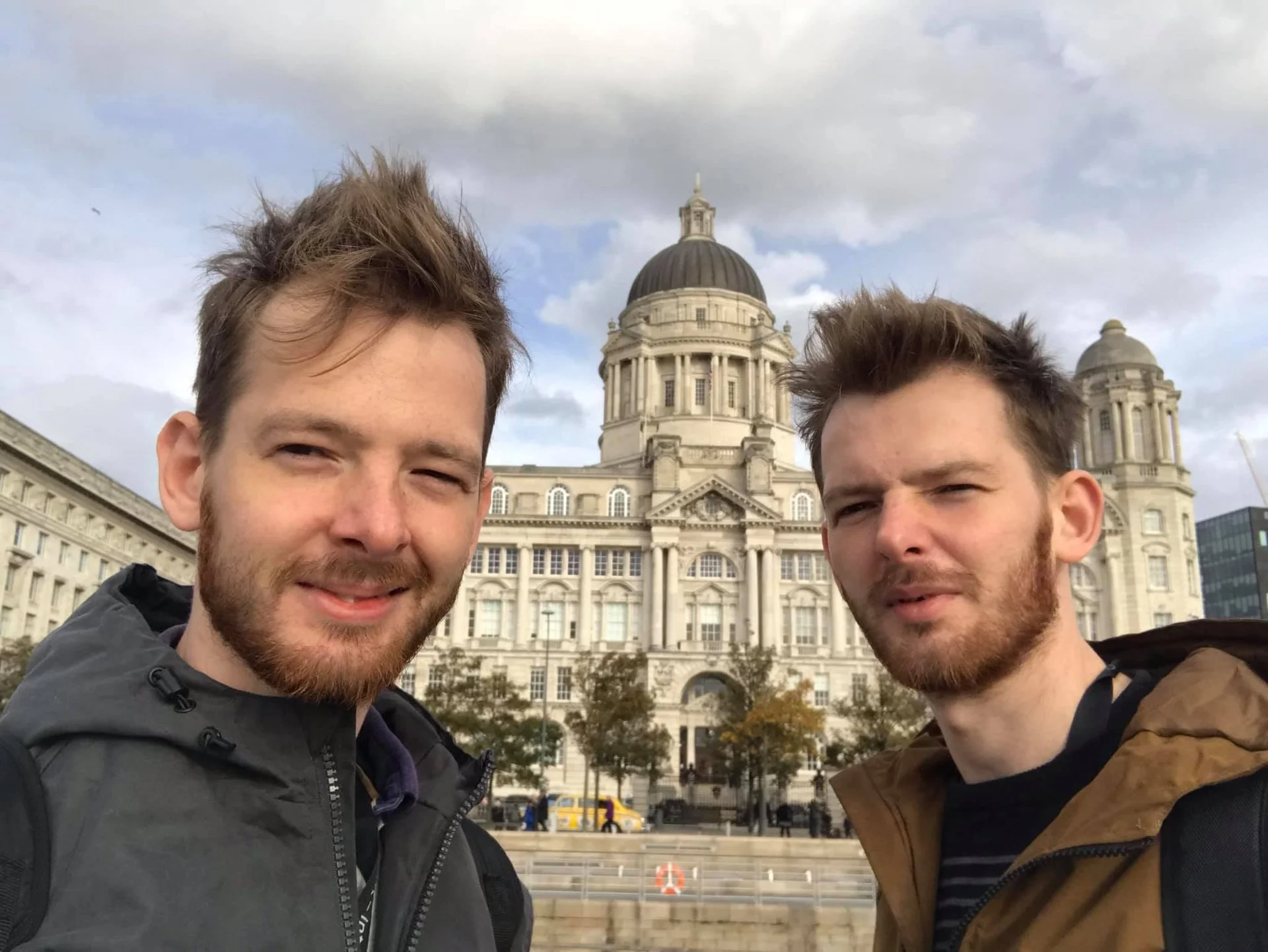 Two young men with beards and tousled hair taking a selfie in front of a historic building with a large dome and two smaller towers, on a cloudy day.