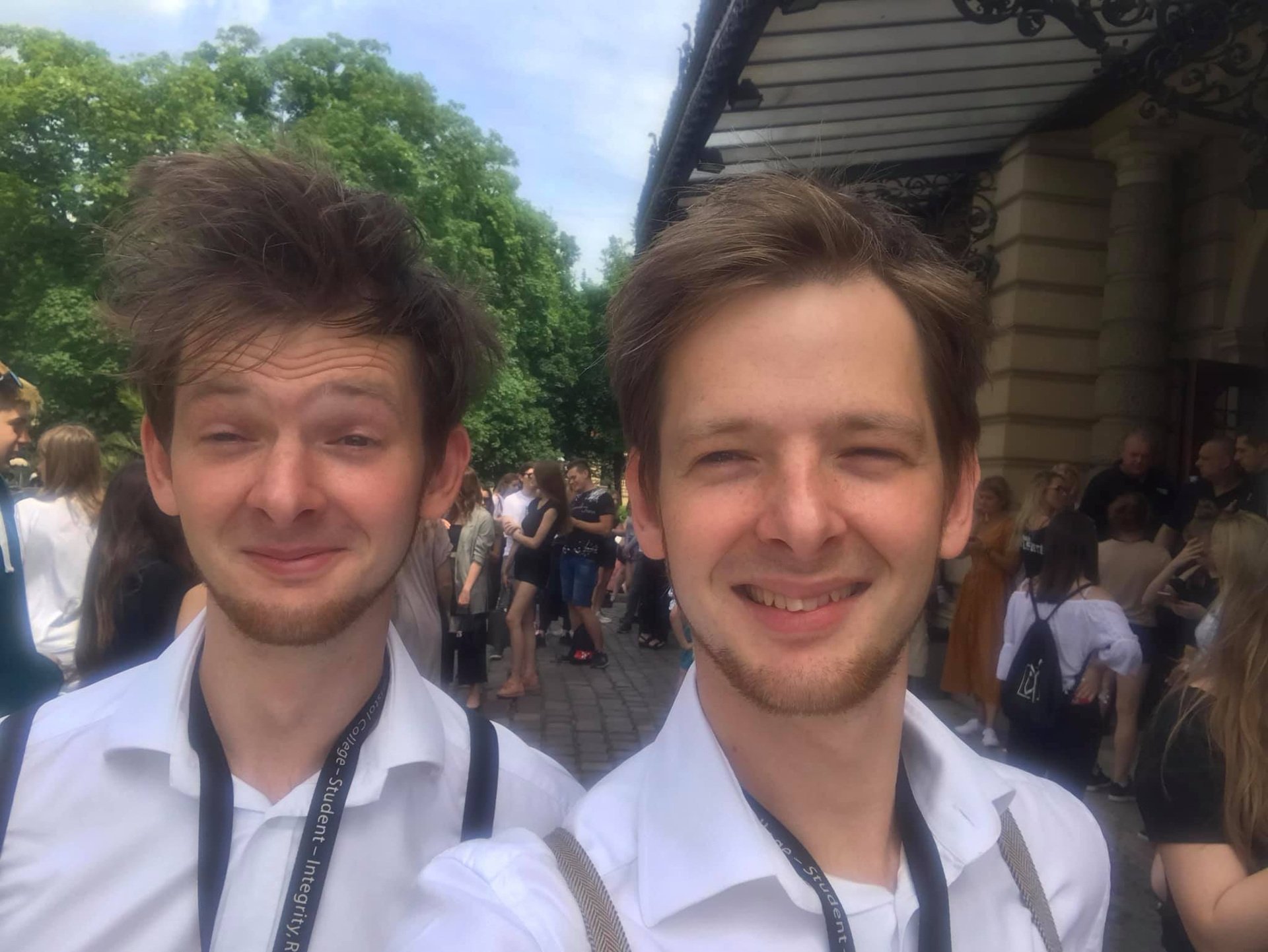 Two young men with light brown hair and beard stubble smiling for a selfie at an outdoor gathering, surrounded by people and green trees.