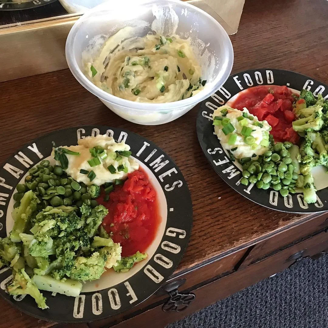 Two plates with steamed broccoli, green peas, mashed potatoes with green onion, and a serving of chopped tomatoes. A bowl of mashed potatoes with green onion is placed above the plates.