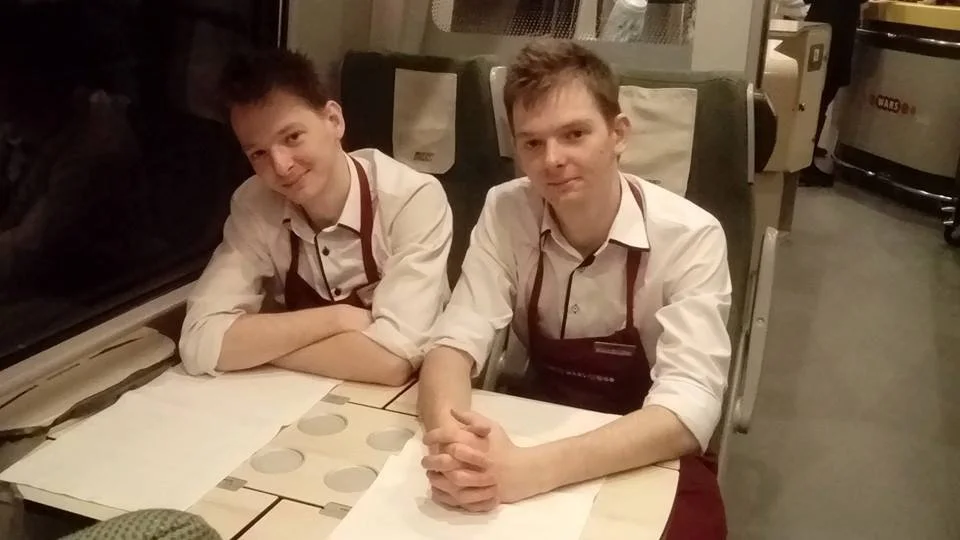 Two young male waiters in white shirts and maroon aprons sitting at a table in a restaurant, smiling at the camera.