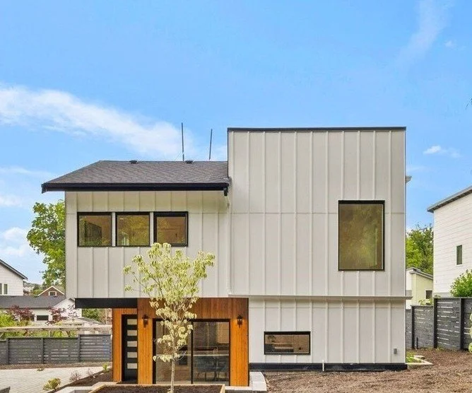Front view of a modern two-story house with vertical white siding, black trim, and wooden accents, featuring large windows and a small tree in the yard.