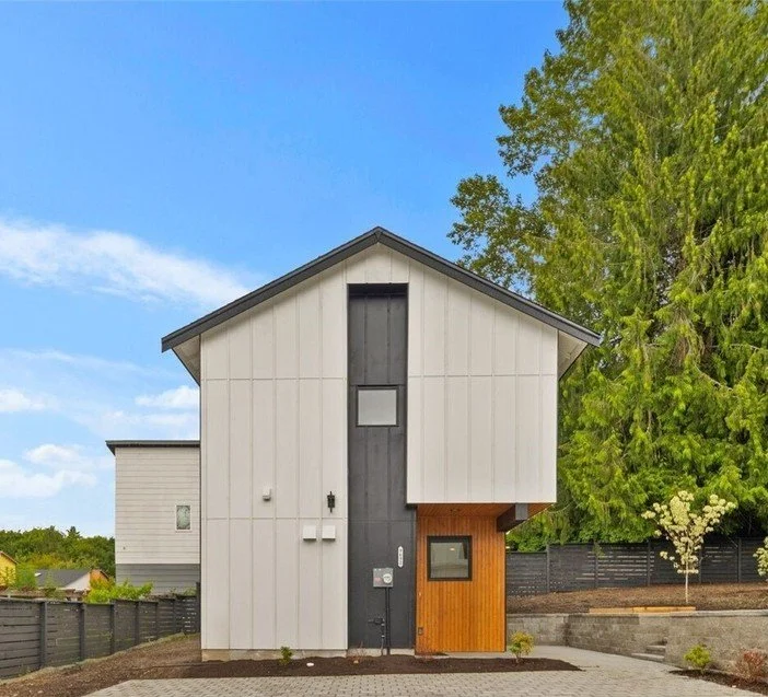 Modern two-story white house with a black front door and a wood-paneled section, surrounded by green trees and a paved driveway.