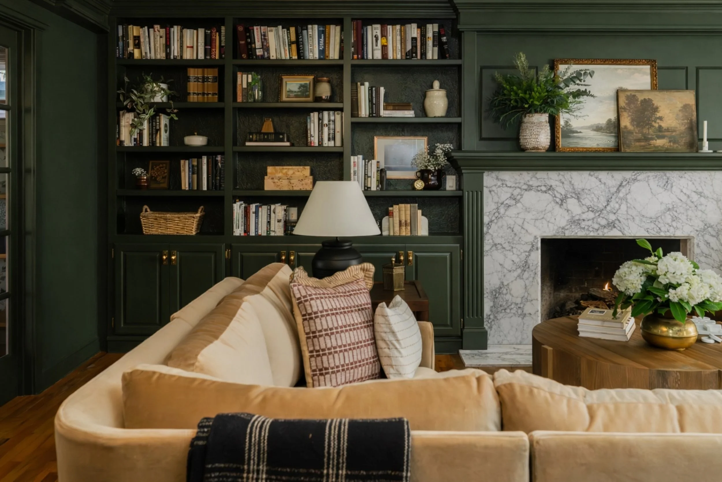 Living room with white built-in shelves, a fireplace with white marble surround, a landscape painting above, and seating with pillows and a plant on a round table.