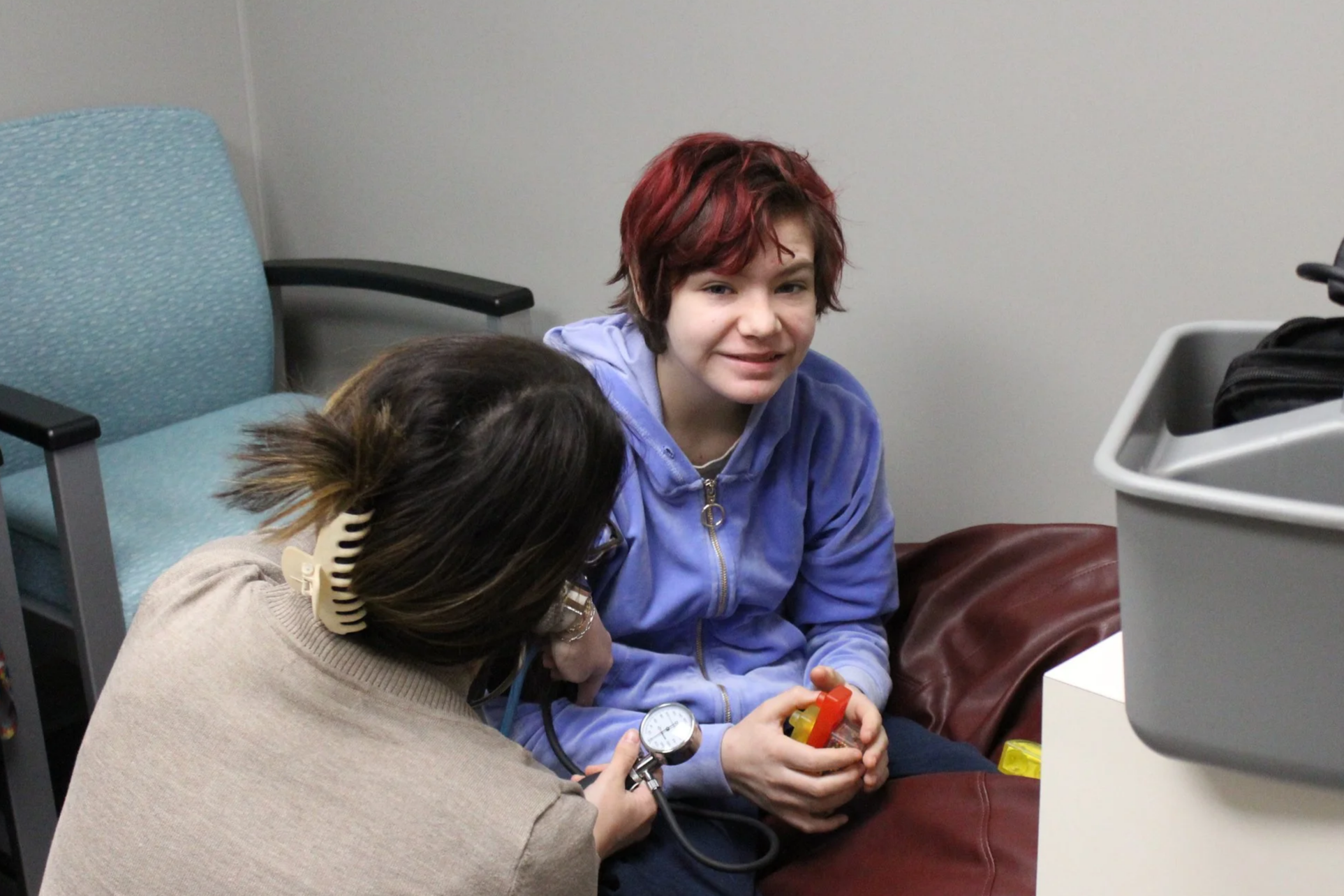 A healthcare professional, wearing blue scrubs and pink long sleeves, is using a stethoscope to examine a young girl with curly blonde hair in a wheelchair, who is talking and has a surprised expression.
