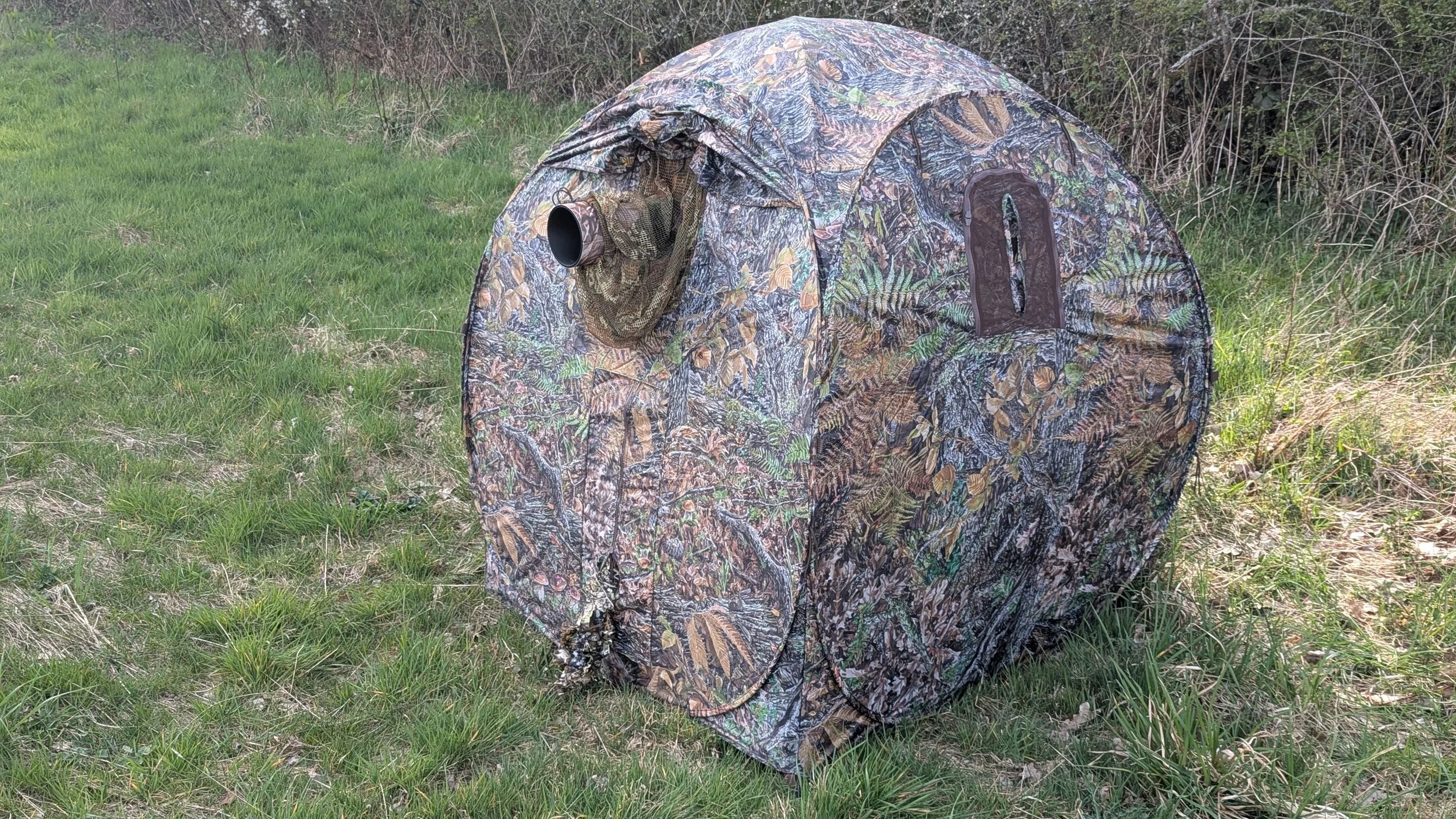 Camouflage hunting blind set up on grassy field with bushes in the background.