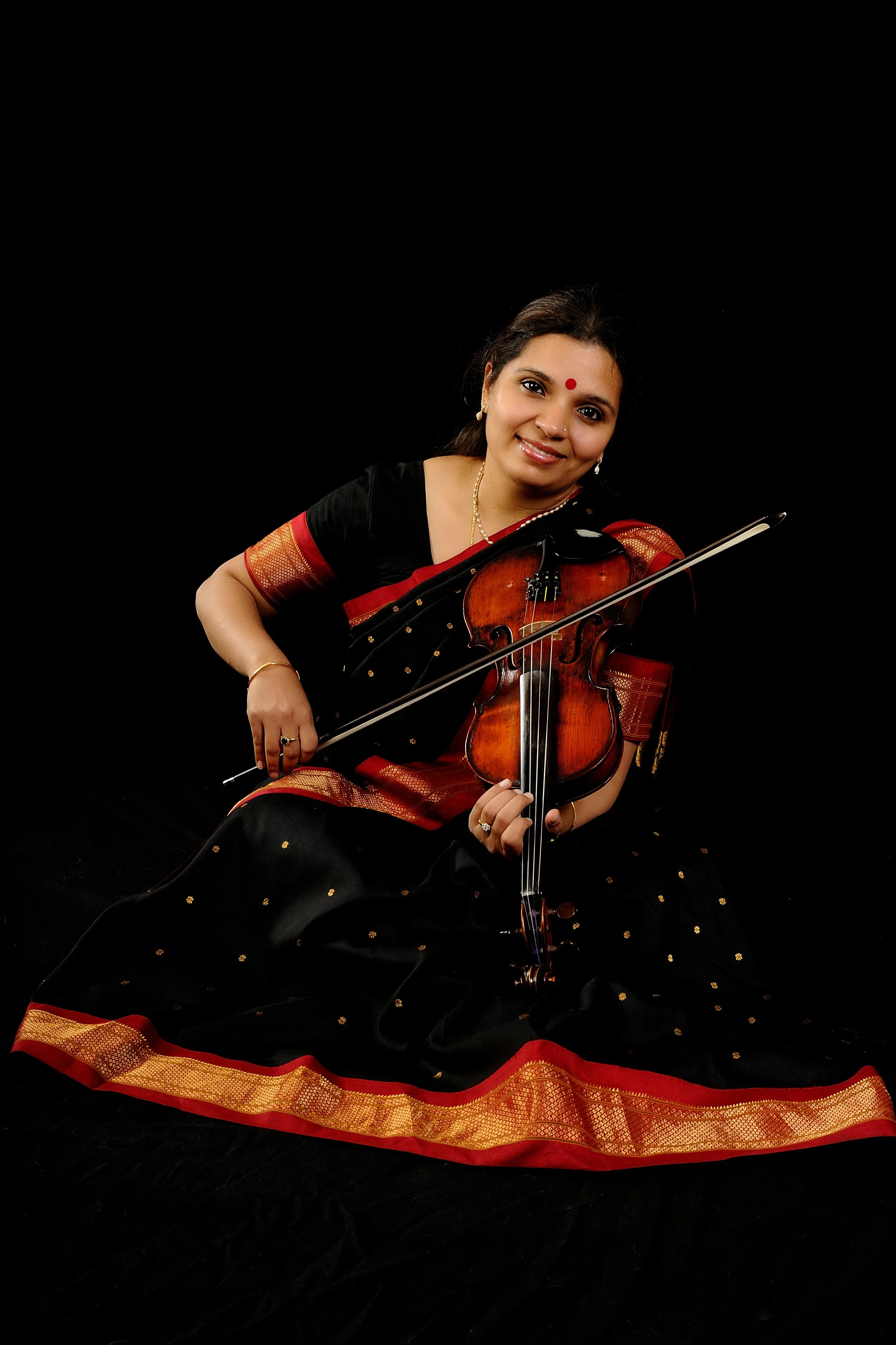 A smiling woman dressed in a black and red traditional saree, sitting on the floor, playing a violin against a black background.