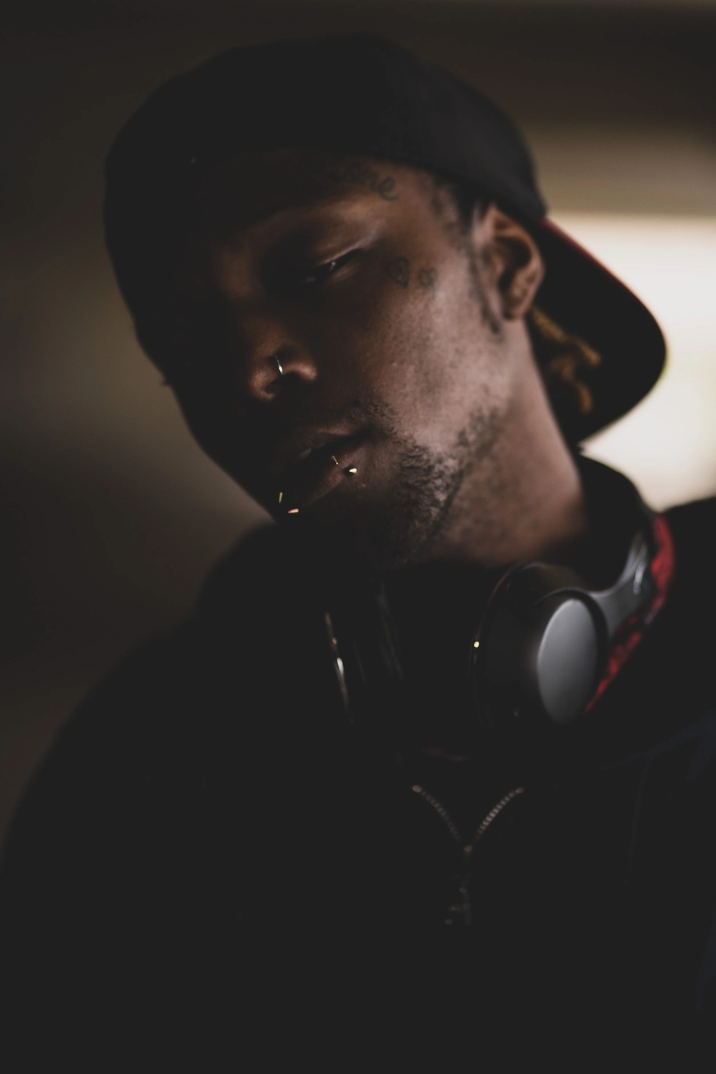 A young man with dark skin wearing a backwards black cap, headphones around his neck, and multiple facial piercings, looking down in a dimly lit room.