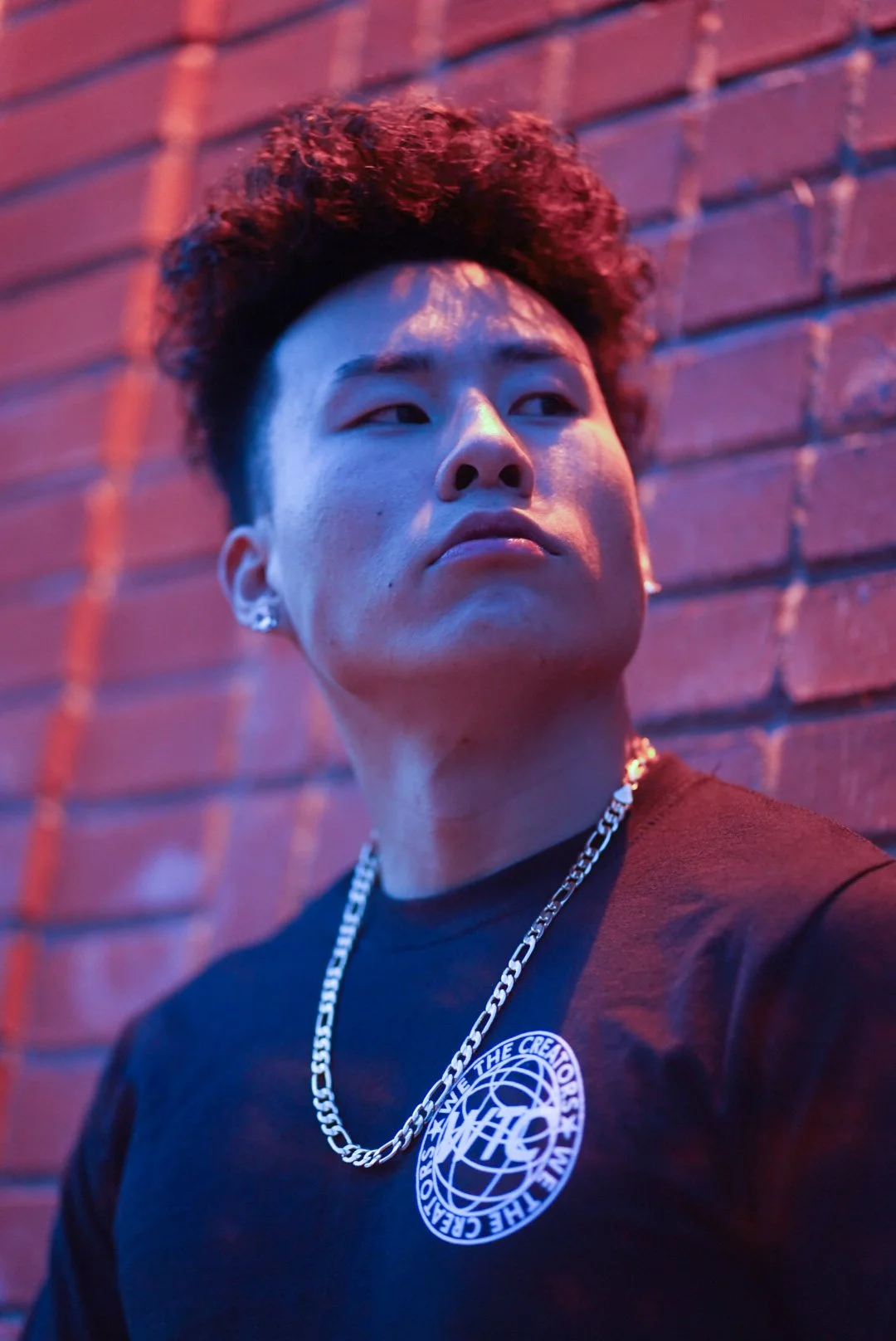 A young man with curly hair and earrings, wearing a black t-shirt with a circular logo and a silver chain, leaning against a brick wall illuminated with pink and blue neon lighting.