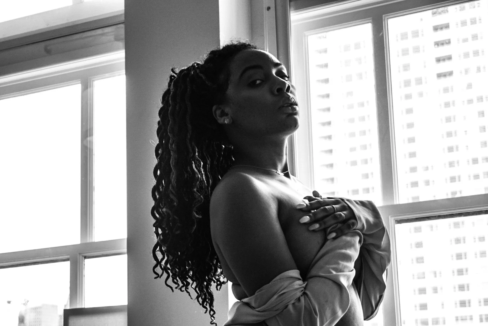 Black and white photograph of a woman with long, curly hair standing near a window, partially covered by an open shirt, looking at the camera with a serious expression.