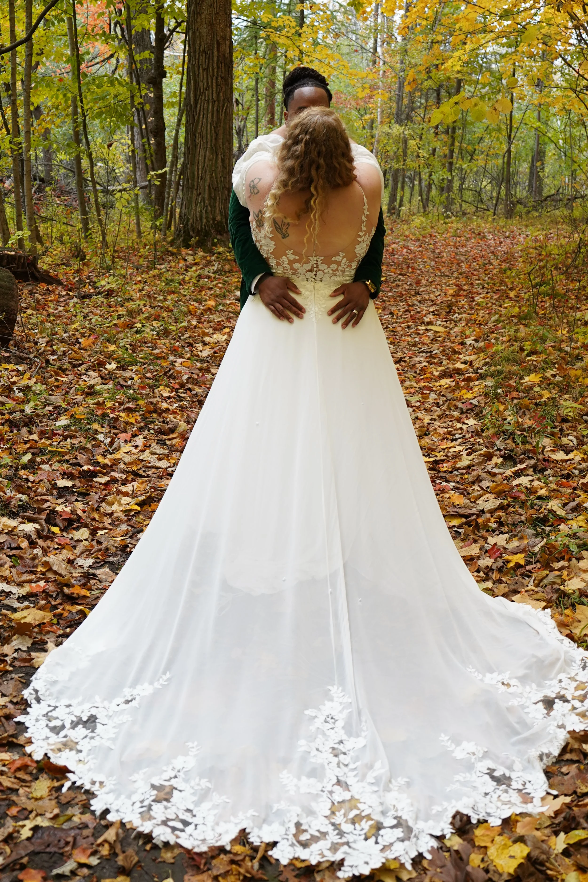A bride and groom embracing on a leaf-covered forest path surrounded by fall foliage.