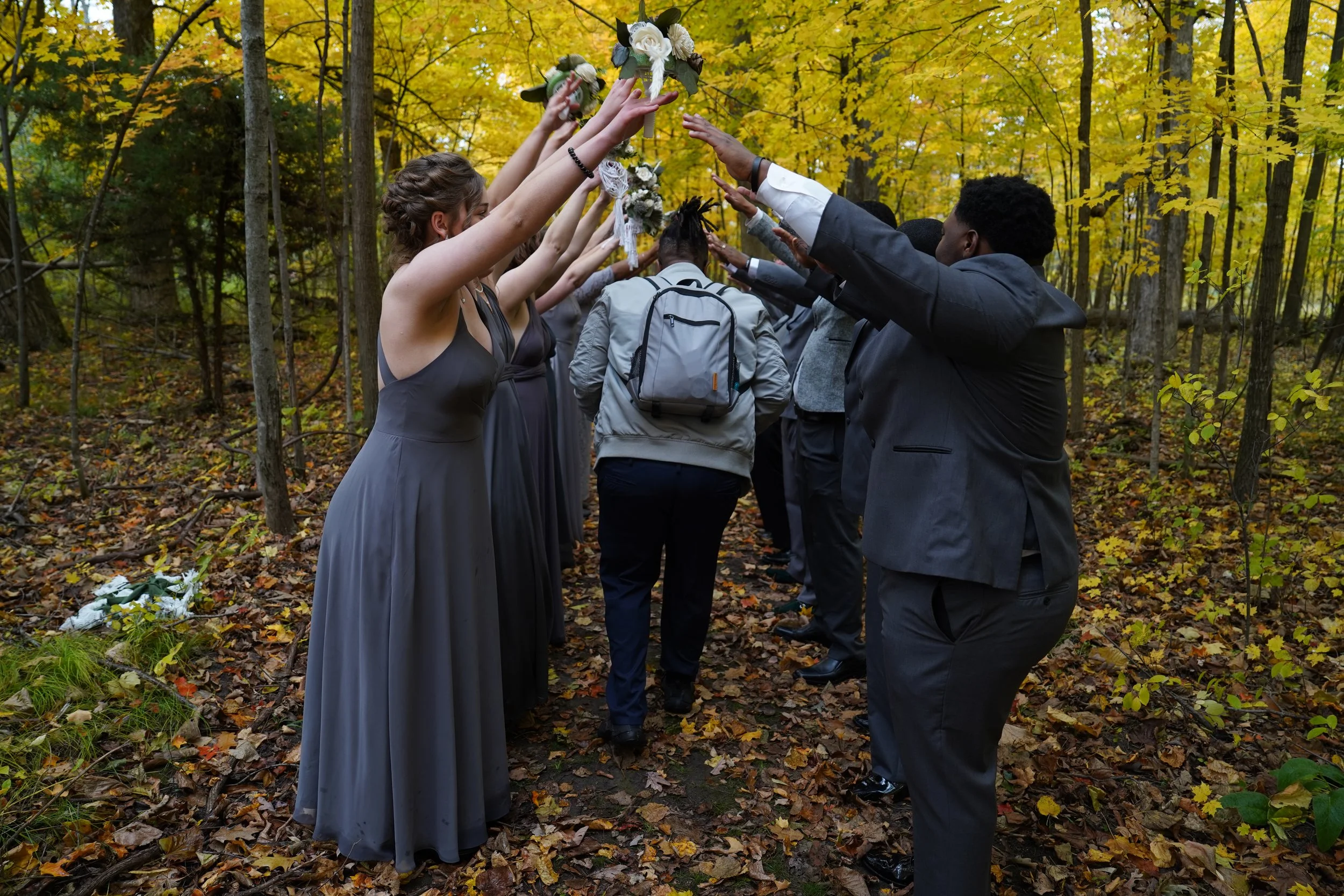 People dressed in formal attire participating in a wedding procession in a forest during autumn, with yellow leaves on the trees and fallen on the ground.