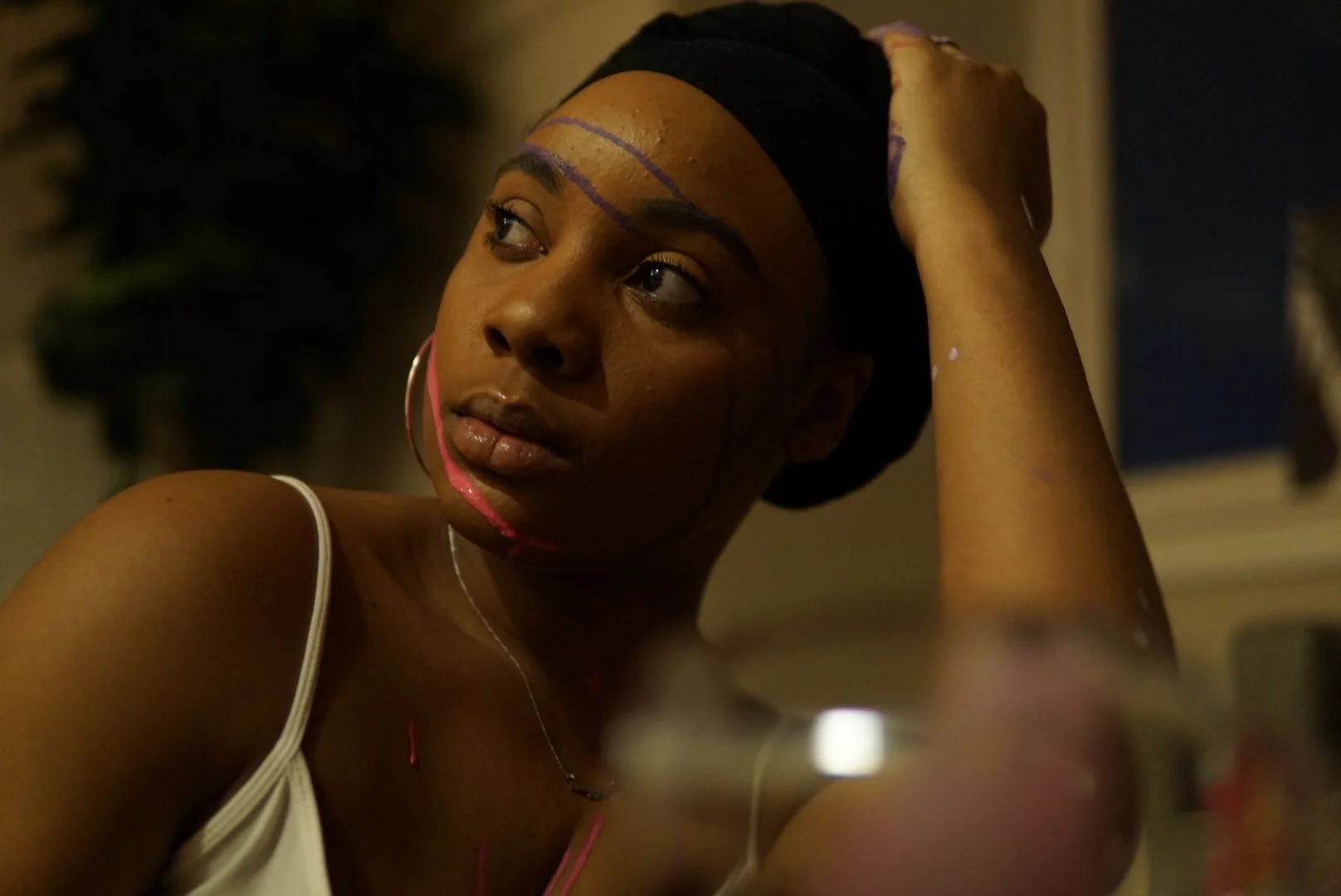 Young woman with dark skin and short hair, wearing a white tank top and large hoop earrings, looking to the side with a thoughtful expression, with purple streaks of paint on her face and arm, and a glass of wine in the foreground.