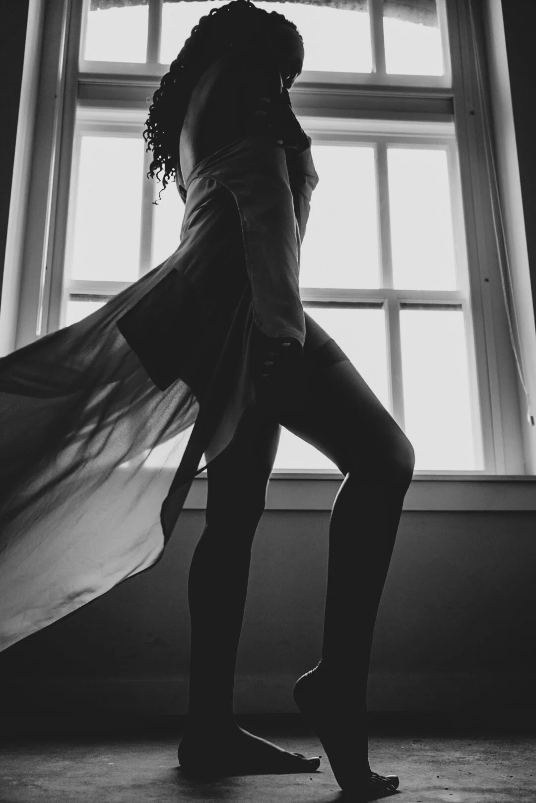Silhouette of a person standing by a window with natural light, wearing a coat with a flowing fabric, in black and white.