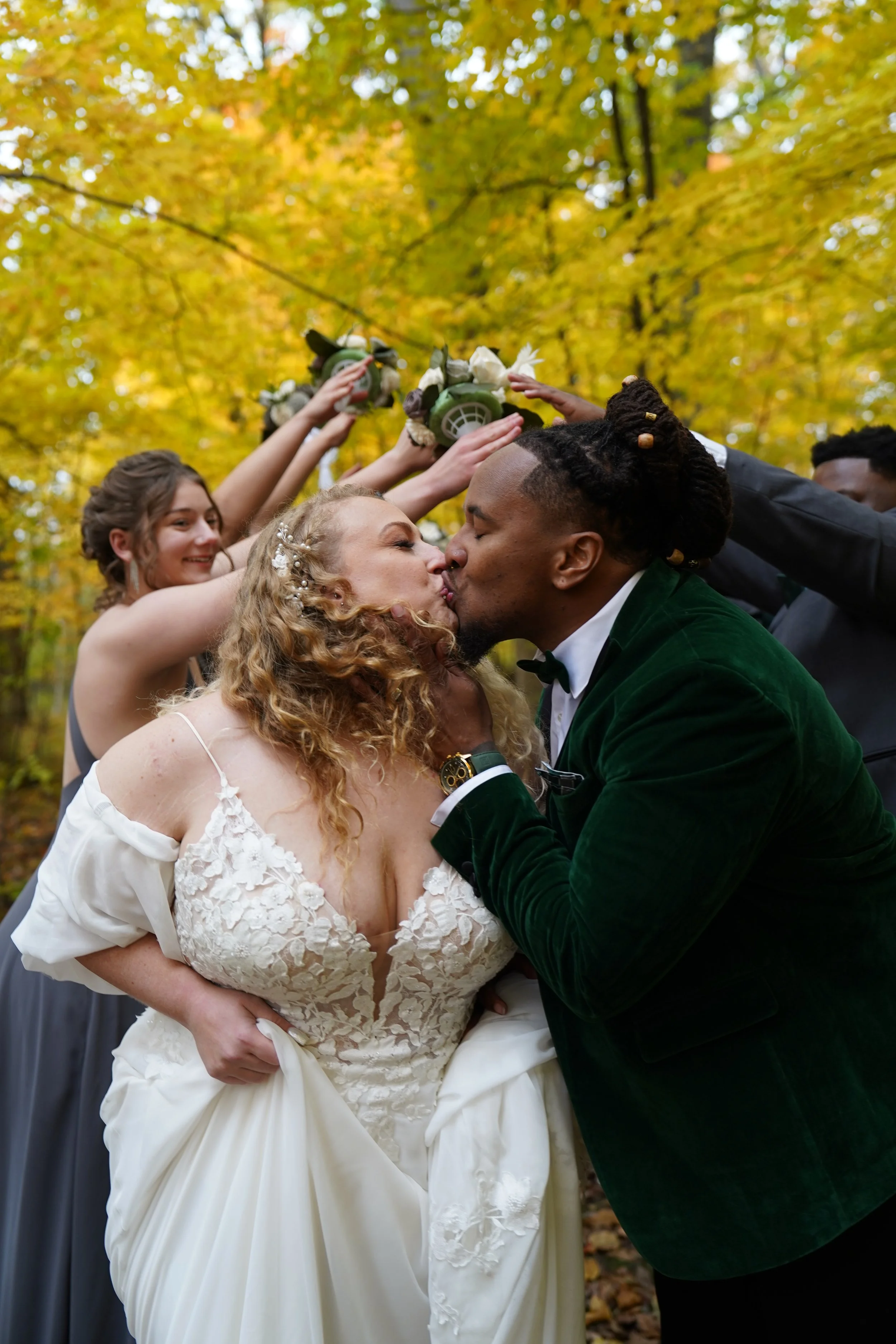A wedding couple, a bride in a white lace dress and a groom in a green velvet jacket, sharing a kiss during an outdoor ceremony surrounded by friends holding floral arrangements over their heads. Fall leaves in the background.