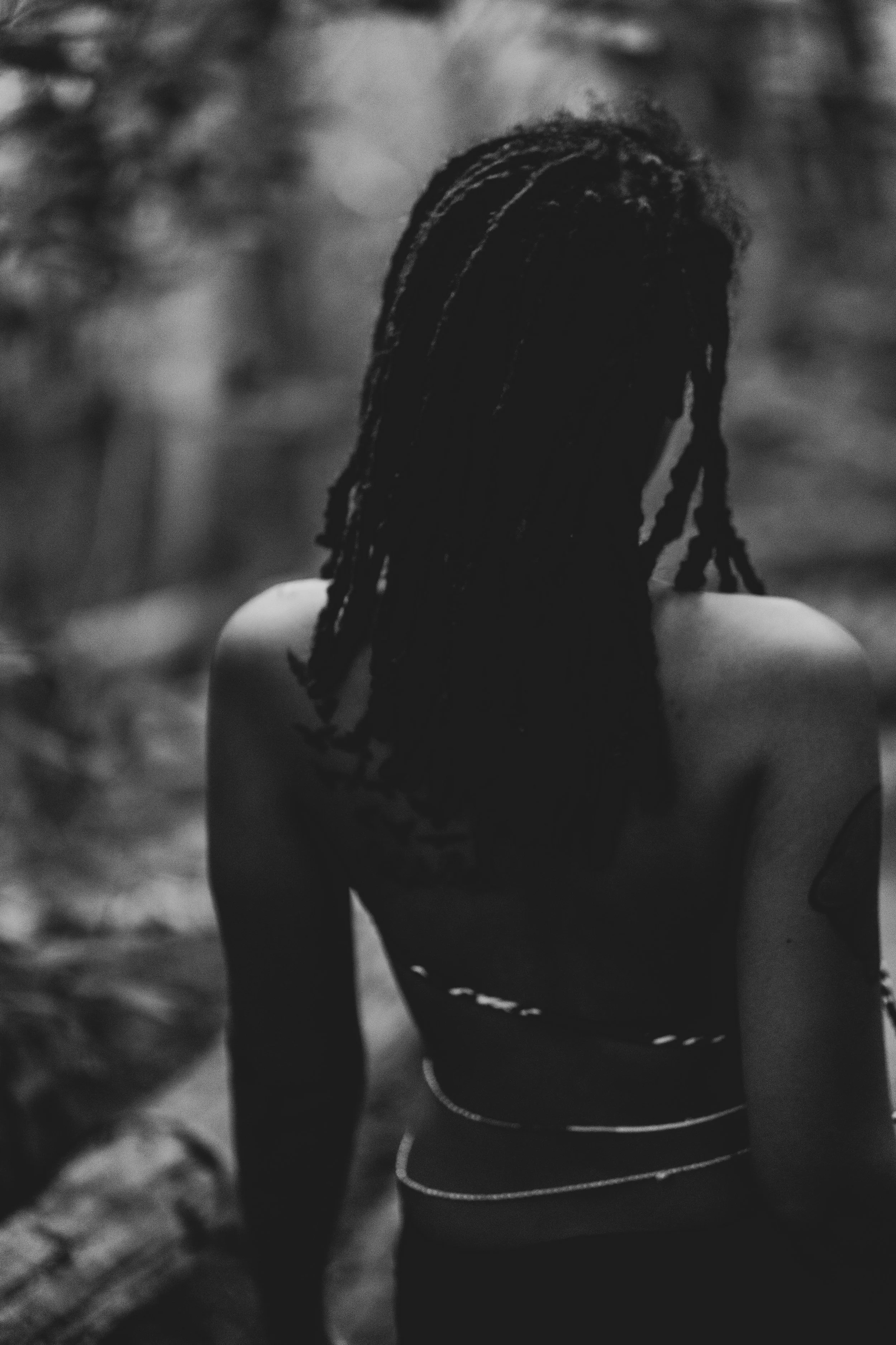 A person with dreadlocks standing outdoors with their back to the camera, wearing a strapless top and jewelry, in black and white.