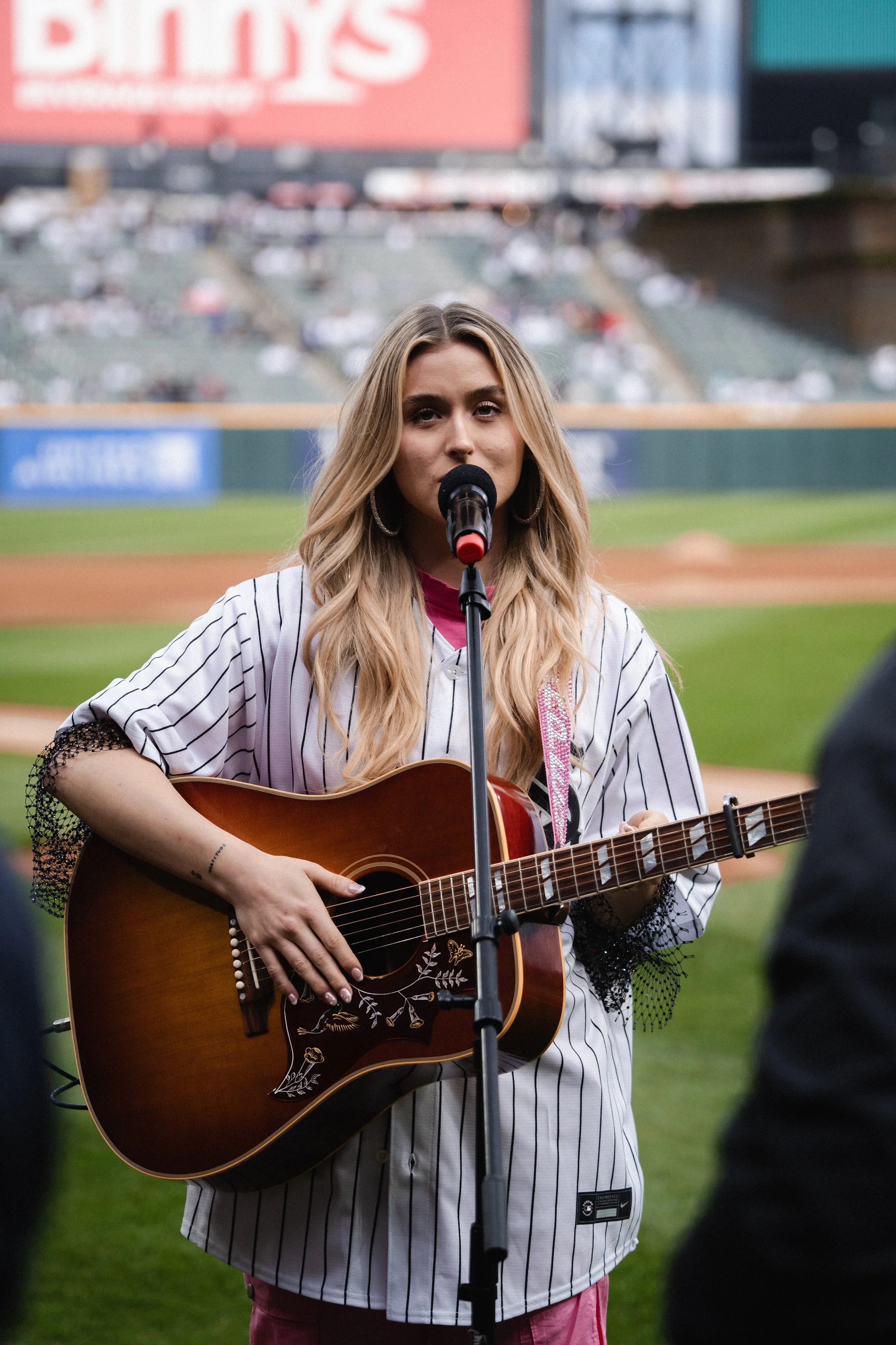 A young woman playing an acoustic guitar and singing into a microphone on a baseball field.