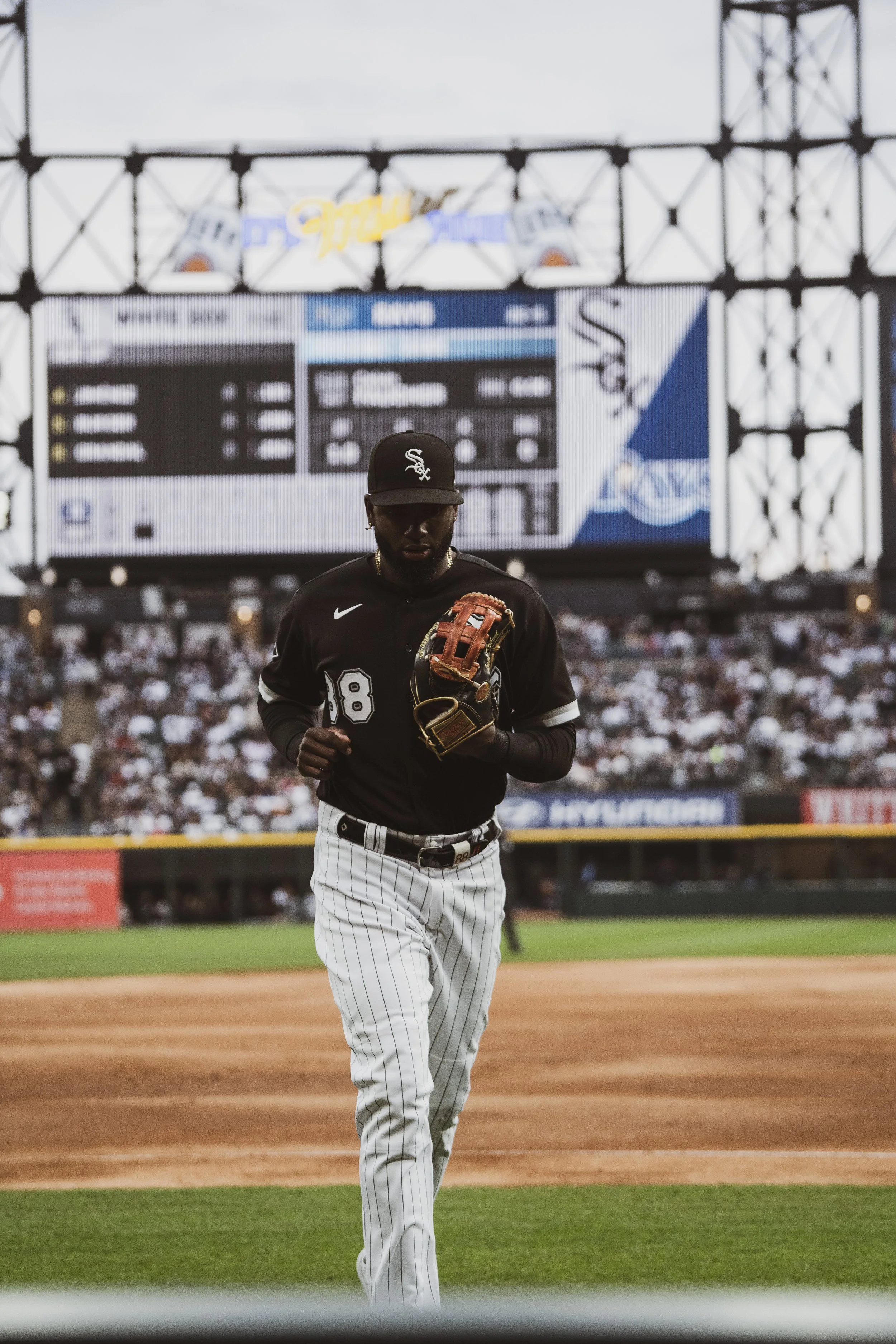 A baseball player in a black uniform and white pinstriped pants running on the field with a glove on his left hand during a game at a stadium, with a large screen and crowd in the background.